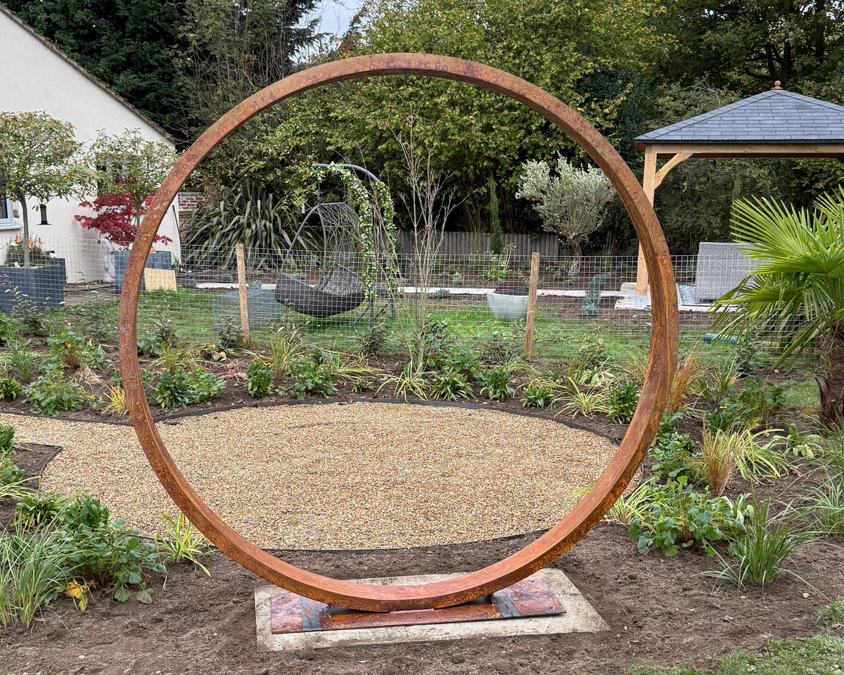 Large Corten steel circular moongate, with a rust patina, a focal point in a garden setting with trees.