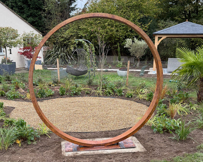 Large Corten steel circular moongate, with a rust patina, a focal point in a garden setting with trees.