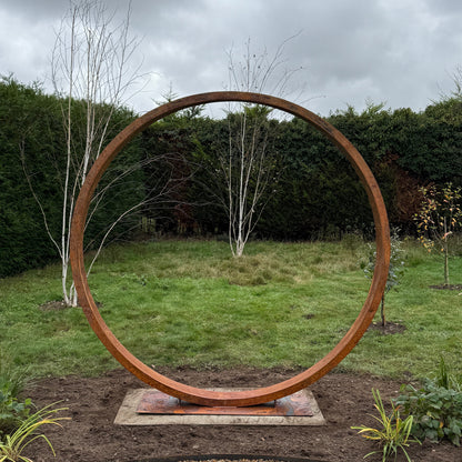 Rust-colored circular Corten steel moongate garden feature,  in a garden setting with a cloudy sky.