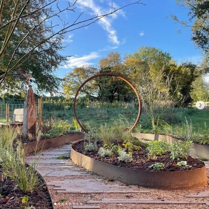 A Corten steel moongate with a view of green grass and trees, in a garden setting with a blue summers sky. 
